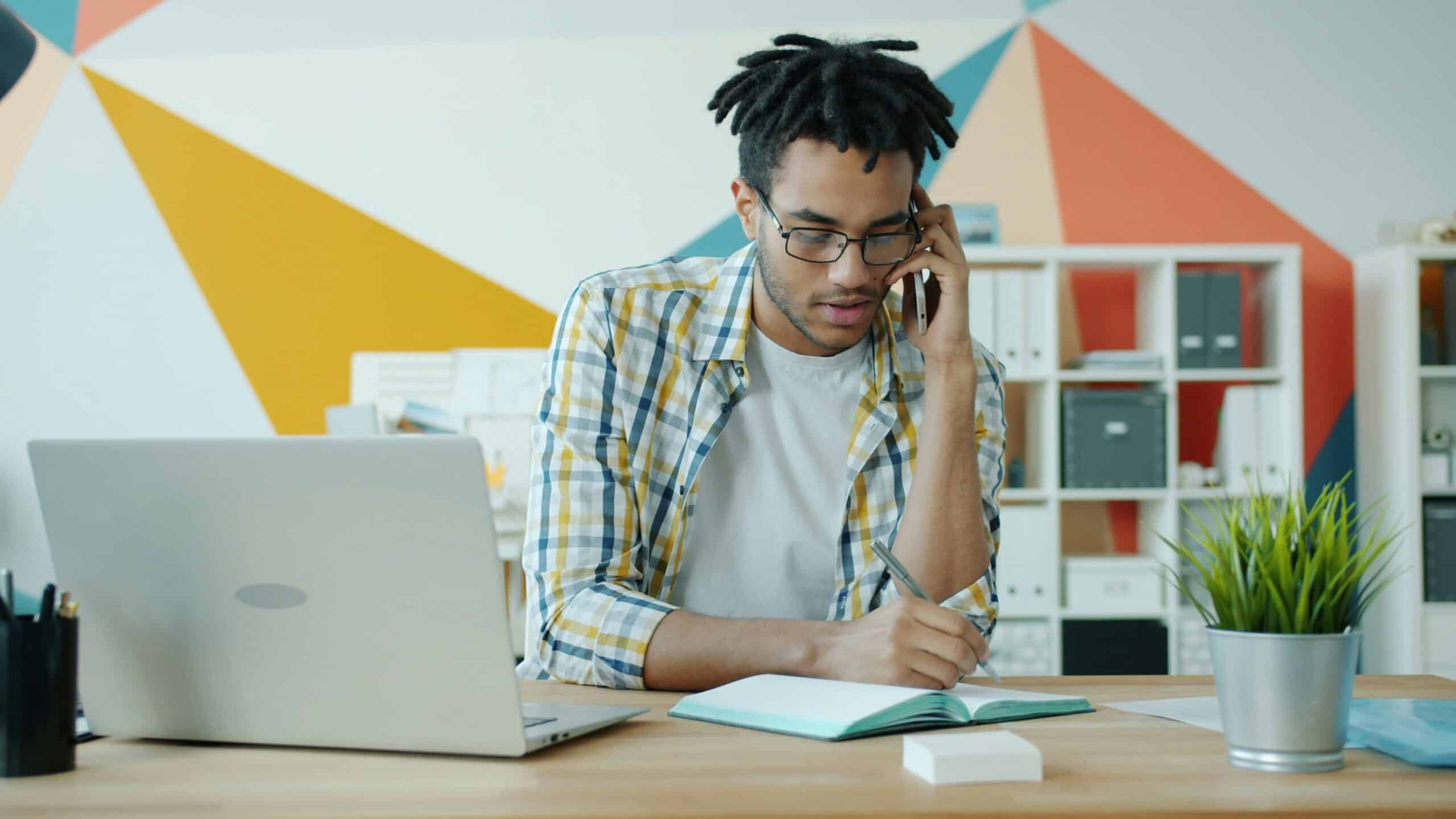 Young professional sitting at a desk with a laptop, writing in a notebook while talking on the phone in a colorful modern office—illustrating executive function and workday organization.