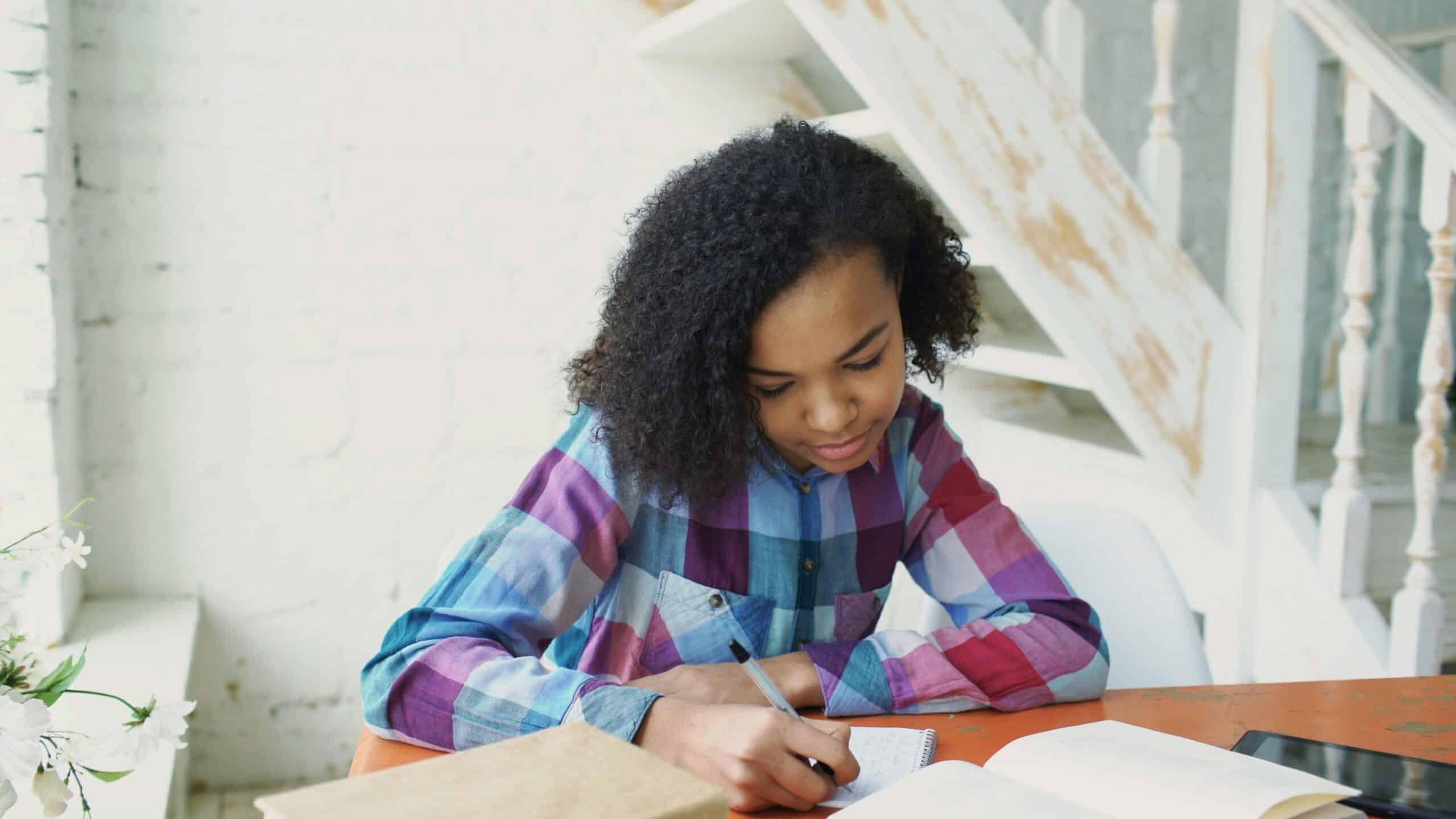 Black woman with curly hair writing in a notebook at a wooden table in a calm, natural-light space, symbolizing reflection, grounding, and somatic trauma recovery work.