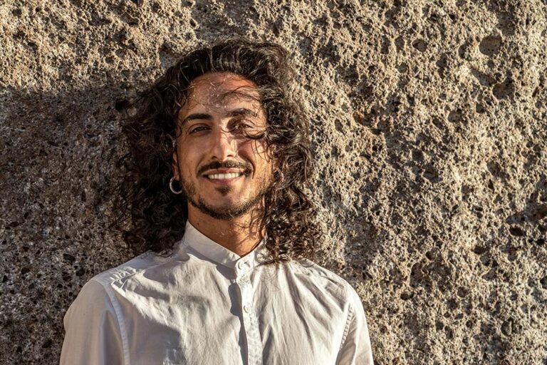 A smiling man with long, curly dark hair and a beard, wearing a white shirt and hoop earrings, leaning against a textured stone wall.