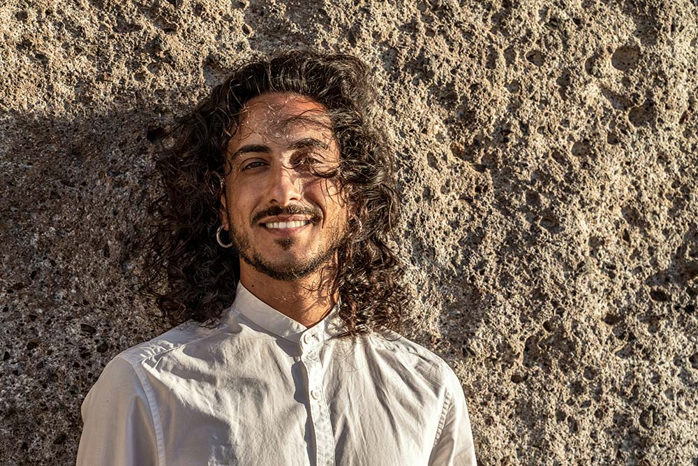 A smiling man with long, curly dark hair and a beard, wearing a white shirt and hoop earrings, leaning against a textured stone wall.