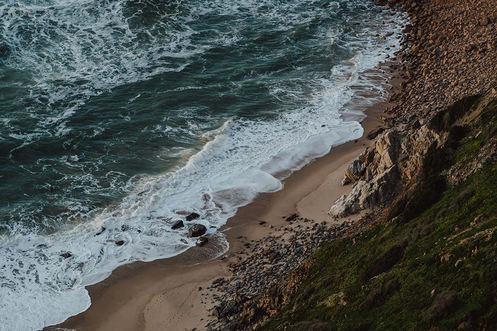 High-angle view of white waves crashing onto a sandy and rocky shoreline at the base of a steep, grassy cliff.