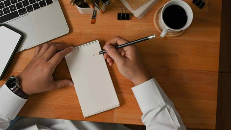 Top view of male hand writing on blank notebook on wooden worktable in home office