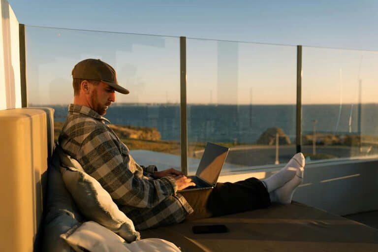 A man working on a laptop on a balcony overlooking the ocean.