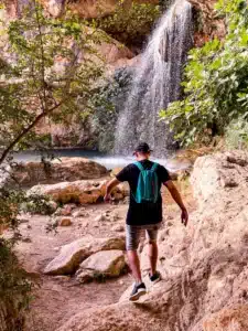 A man with a backpack walking over rocks toward a waterfall in a canyon.