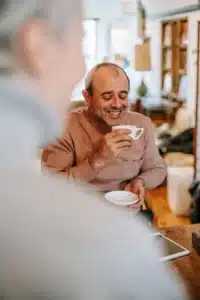 A man smiling while holding a white coffee cup and saucer at a table.