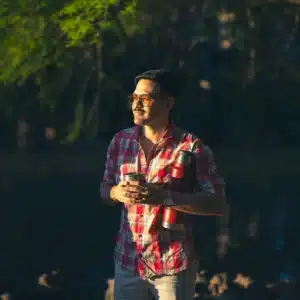 A man wearing sunglasses and a red plaid shirt holding a yerba mate gourd and thermos outdoors.