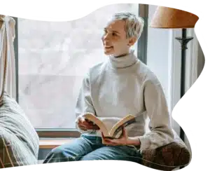 A smiling woman with short hair sitting by a window holding an open book.