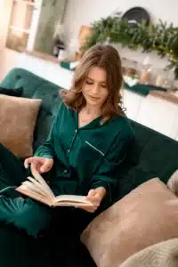 A woman wearing green silk pajamas sitting on a velvet couch and reading a book.