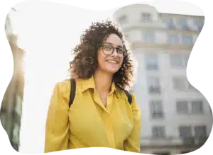 A smiling woman with curly hair and glasses in a yellow shirt outdoors in the city.